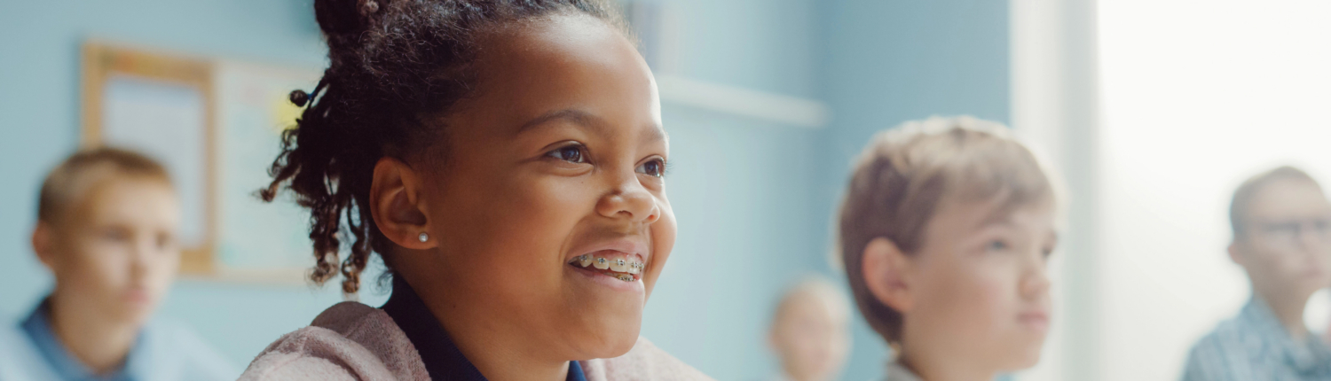 In Elementary School Class: Portrait of a Brilliant Black Girl with Braces Smiles, Writes in Exercise Notebook. Junior Classroom with Diverse Group of Children Learning New Stuff